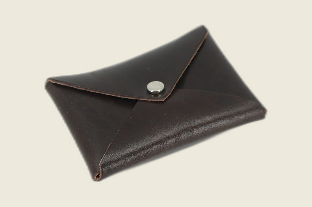 Aegis envelope card wallet in glossy Shinki dark brown glazed horsebutt with nickel snap, closed on a cream backdrop
